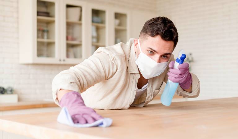 Man in clean shirt wearing a mask holding a bottle in his hand and cleaning a table with a cloth.