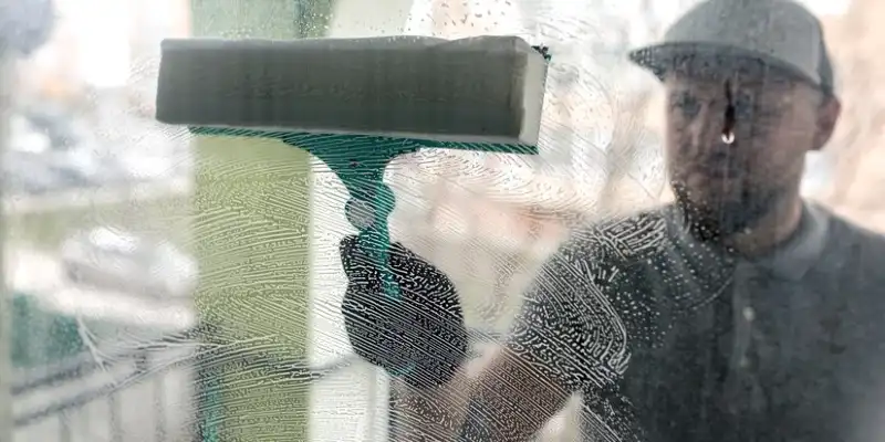 Man in uniform and cap cleaning window with a brush.