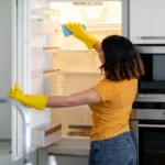 Young housewife wiping shelves in empty fridge while doing cleaning in kitchen