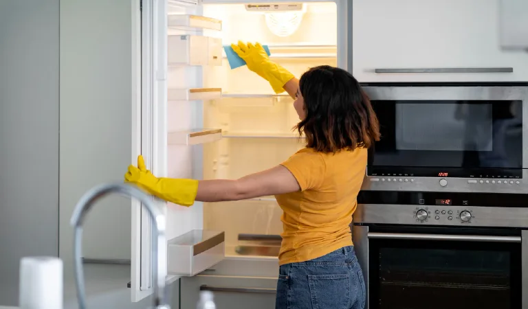 Young housewife wiping shelves in empty fridge while doing cleaning in kitchen