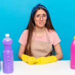 Young pretty house keeper looking puzzled and confused and cleaning products on table