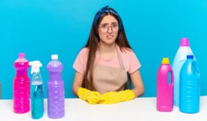 Young pretty house keeper looking puzzled and confused and cleaning products on table