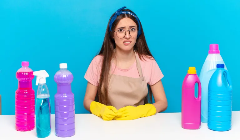 Young pretty house keeper looking puzzled and confused and cleaning products on table